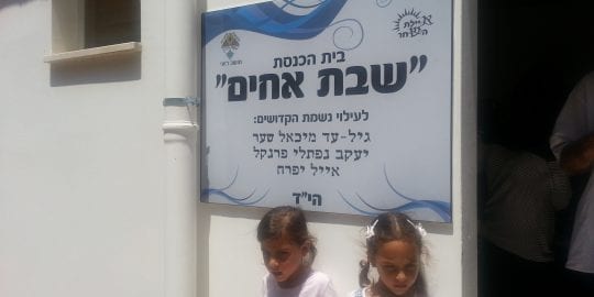 Children from Roi stand in front of the plaque which reads, in Hebrew: the Shevet Achim Synagogue, Dedicated in memory of Gil-Ad Shayar, Naftali Frenkel and Eyal Yifrach.